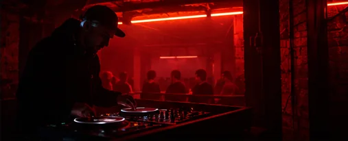 A DJ under red strip lights in a low ceilinged room